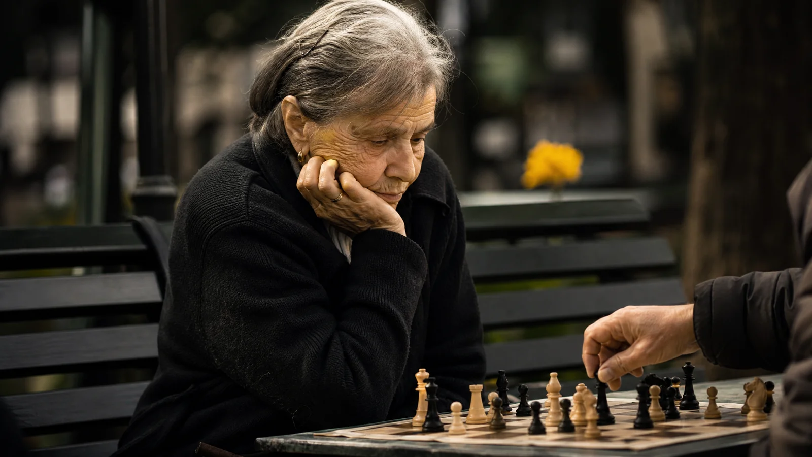 An elderly woman playing chess in a park, studying the board with her chin on her hand.