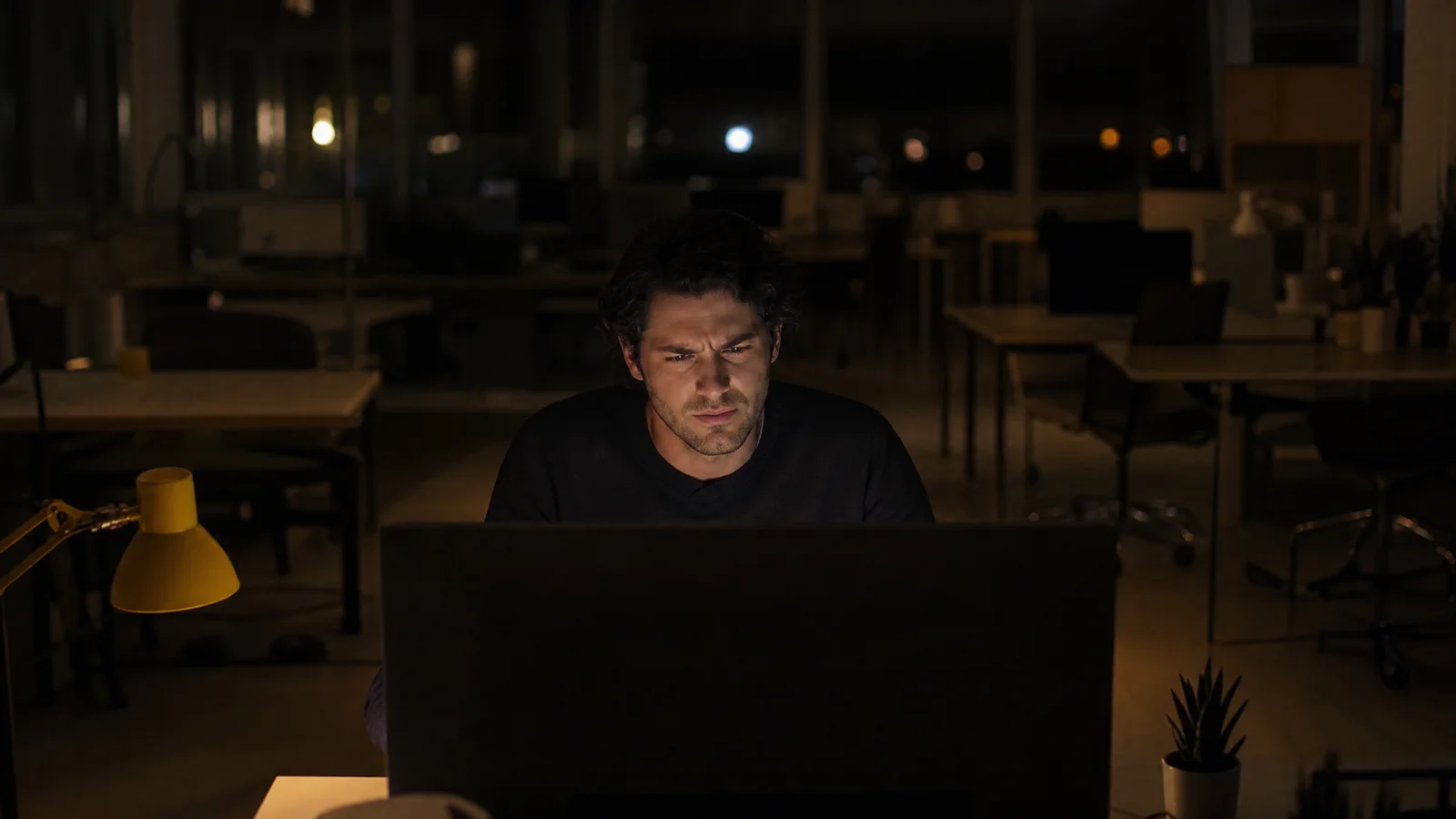 A man sits alone in a dark office at night, his face lit by a monitor, looking puzzled at the screen.